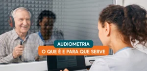 Idoso dentro de cabine acústica com fones de ouvido sinalizando percepção de som durante exame de audiometria realizado por fonoaudióloga.